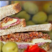 deli sandwich topped with muffaletta, next to 2 olives some roasted red peppers and a jar of olives