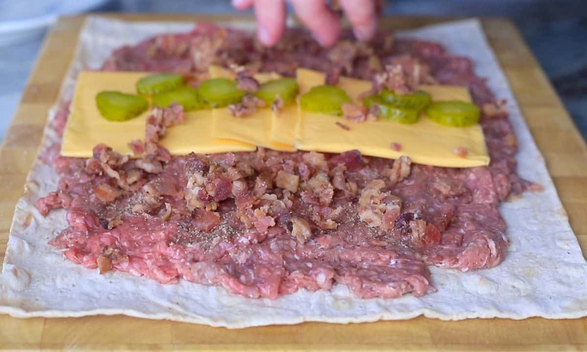 adding bacon bits onto the lavash bread also topped with ground meet, cheese slices and pickels.