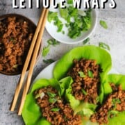 three chicken lettuce wraps on a white dish with a bowl of chicken, two chopsticks and a side dish with sliced green onions.