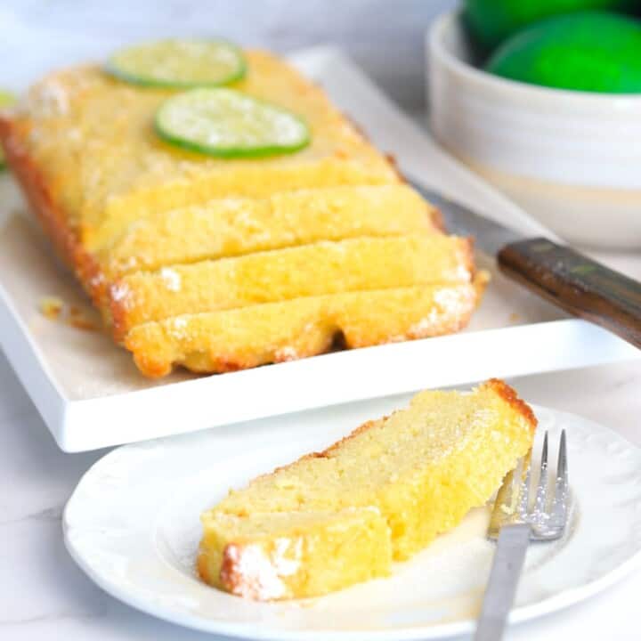 Lime drizzle cake on a white place cut into slices with another plate in front with a slice and a fork and a bowl of limes in the background.