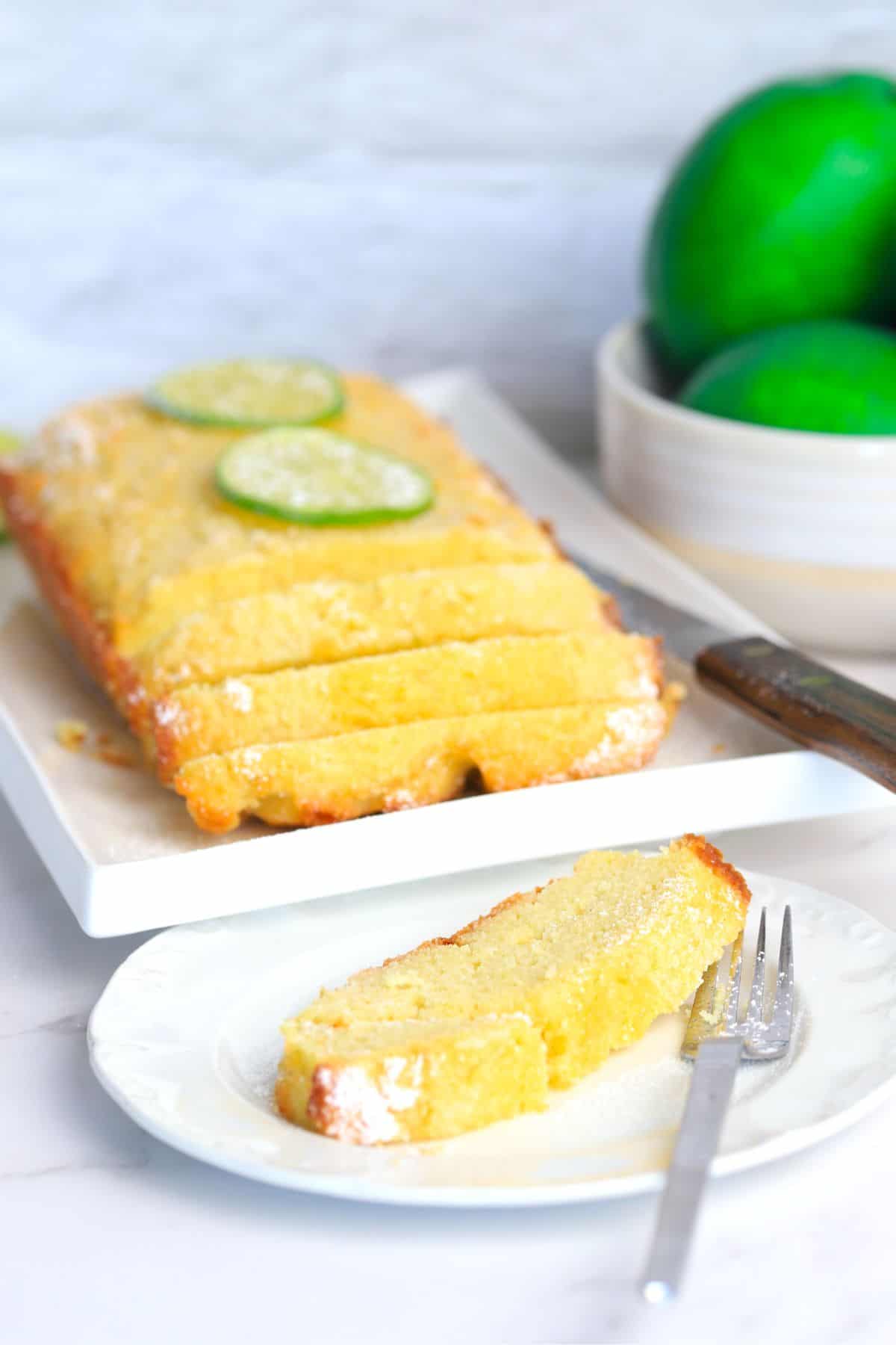 Lime drizzle cake on a white place cut into slices with another plate in front with a slice and a fork and a bowl of limes in the background.