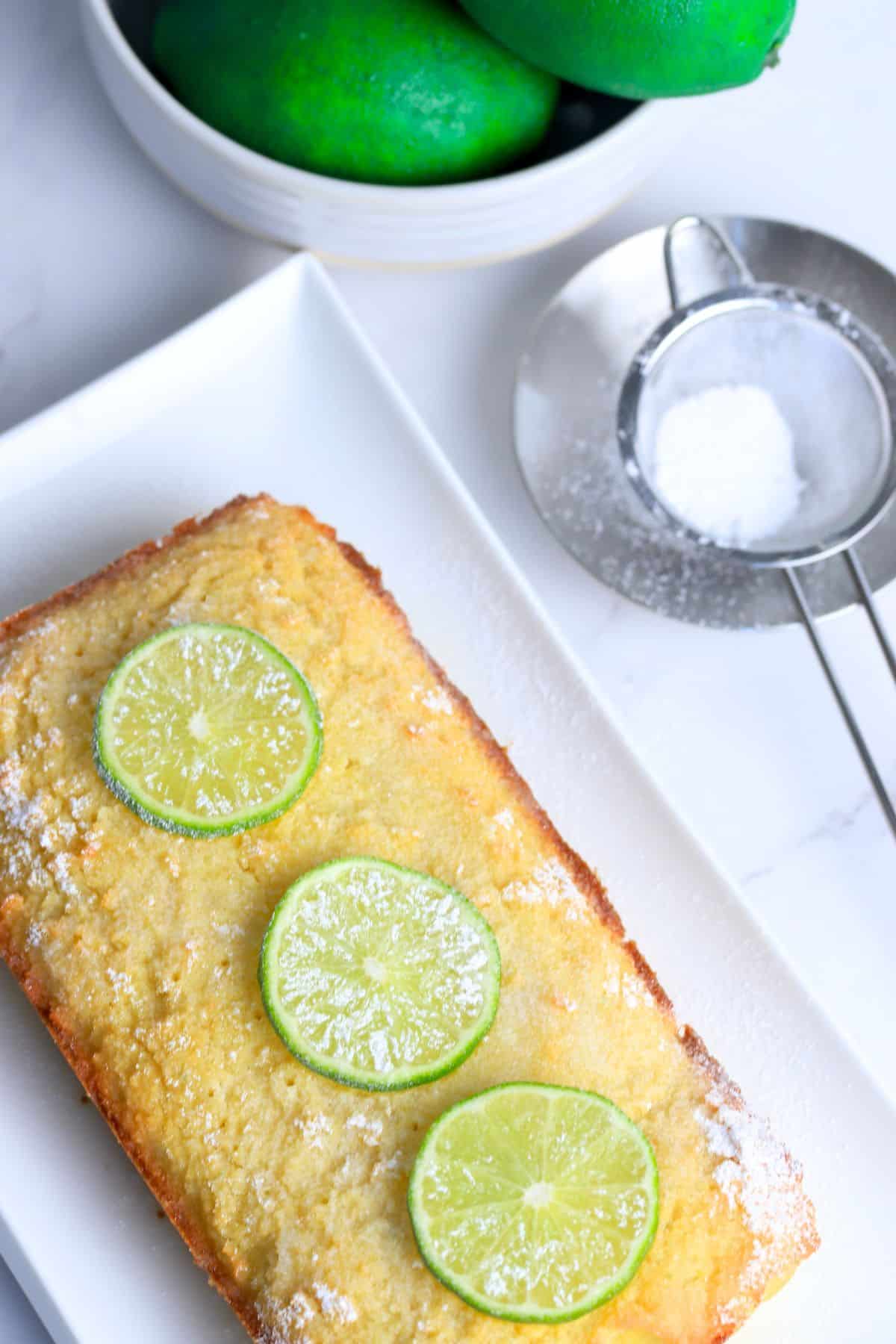 Lime drizzle cake loaf topped with a sprinkle of powdered sugar and 3 lime slices, next to a strainer with powedered sugar and a bowl with limes.