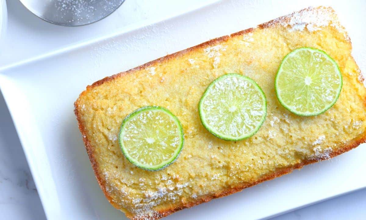 Lime drizzle cake loaf topped with a sprinkle of powdered sugar and 3 lime slices.