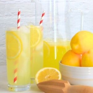 two glasses filled with lemonade next to a half filled jug, a bowl of lemons, a half cut lemon and a reamer.