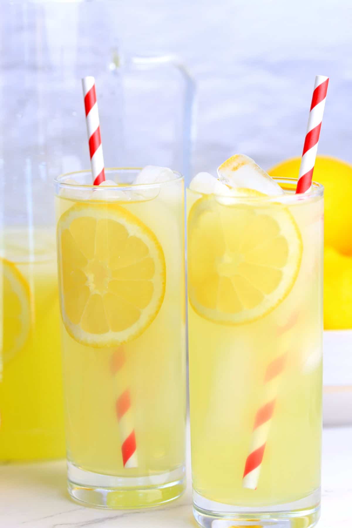 two glasses filled with lemonade next to a half filled jug, a bowl of lemons.