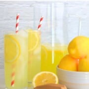 two glasses filled with lemonade next to a half filled jug, a bowl of lemons, a half cut lemon and a reamer.