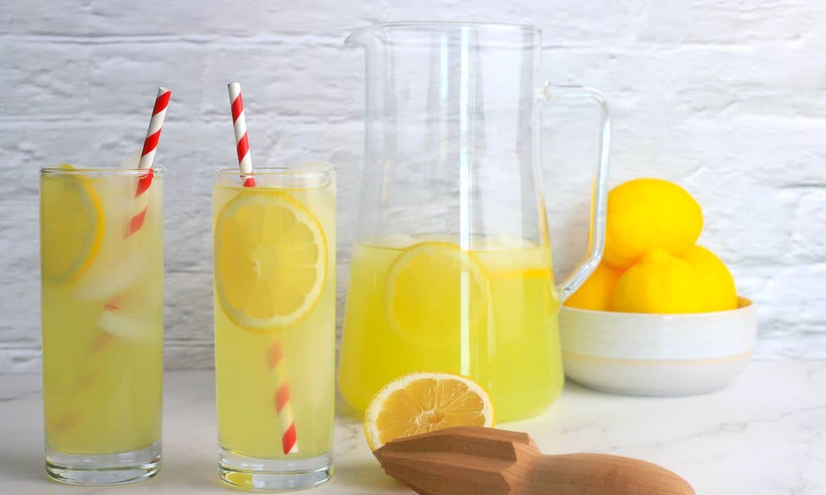 two glasses filled with lemonade next to a half filled jug, a bowl of lemons, a half cut lemon and a reamer.