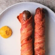 two bacon-wrapped hot dog on a plate with a dollop of mustard.