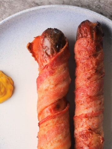 two bacon-wrapped hot dog on a plate with a dollop of mustard.