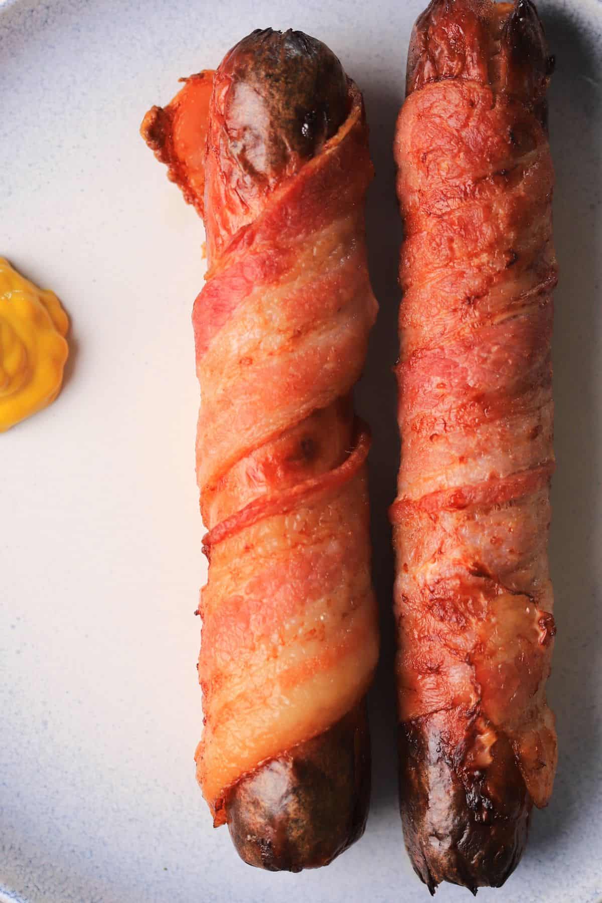 two bacon-wrapped hot dogs on a plate with a dollop of mustard.