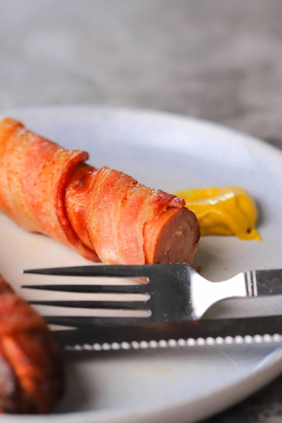 bacon-wrapped hot dog on a plate with a fork and a dollop of mustard.
