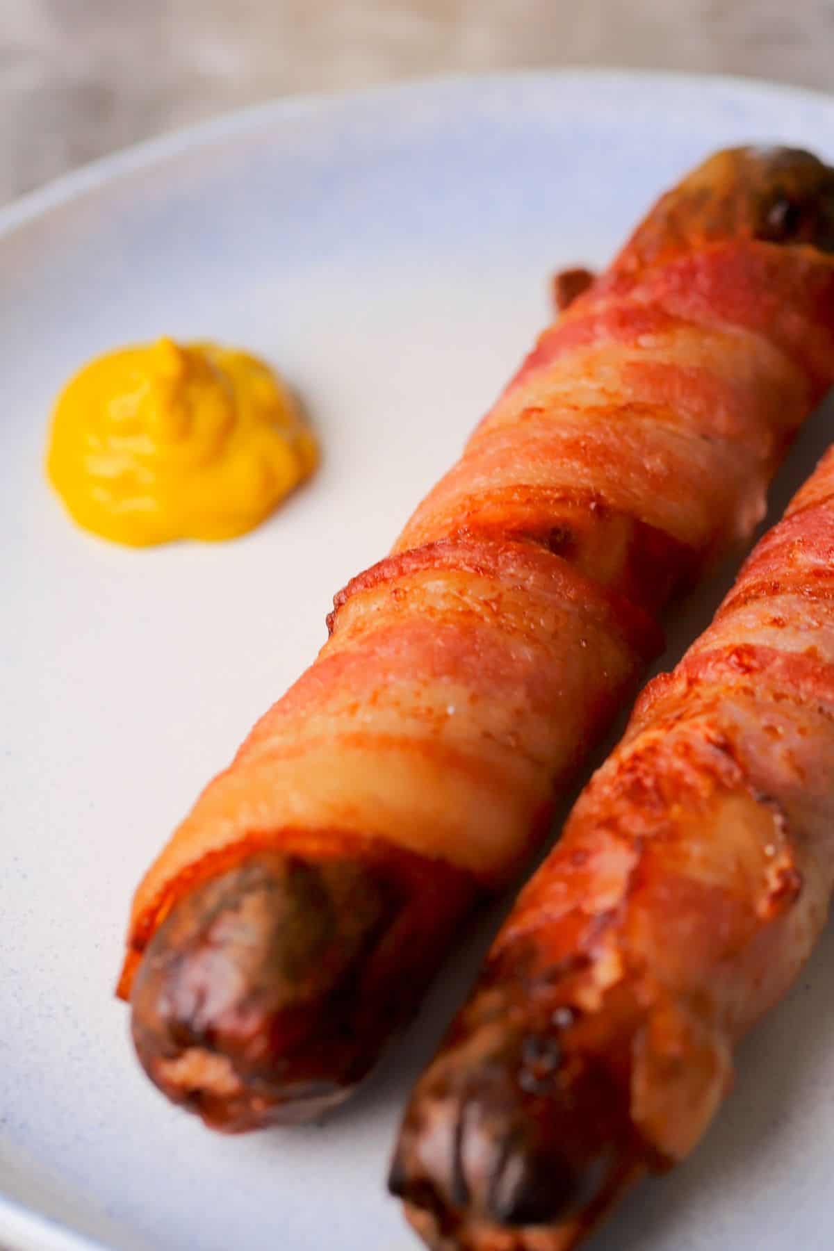 two bacon-wrapped hot dogs on a plate with a dollop of mustard.