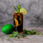 a black mojito in a tall glass garnished with mint and lime with a full lime next to the glass.