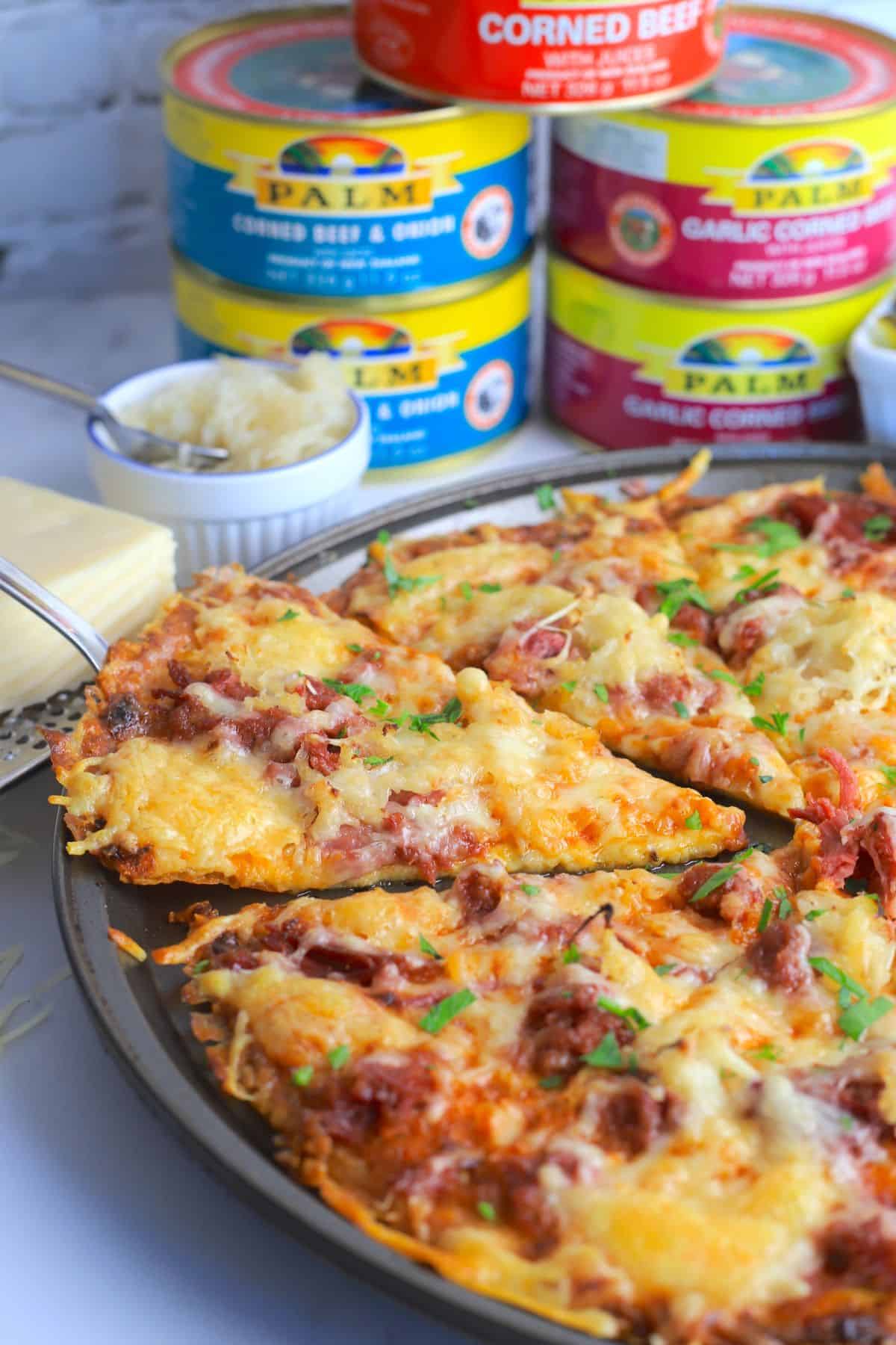 Reuben pizza next to sauerkraut and cans of corned beef and a bowl with sliced pickles.