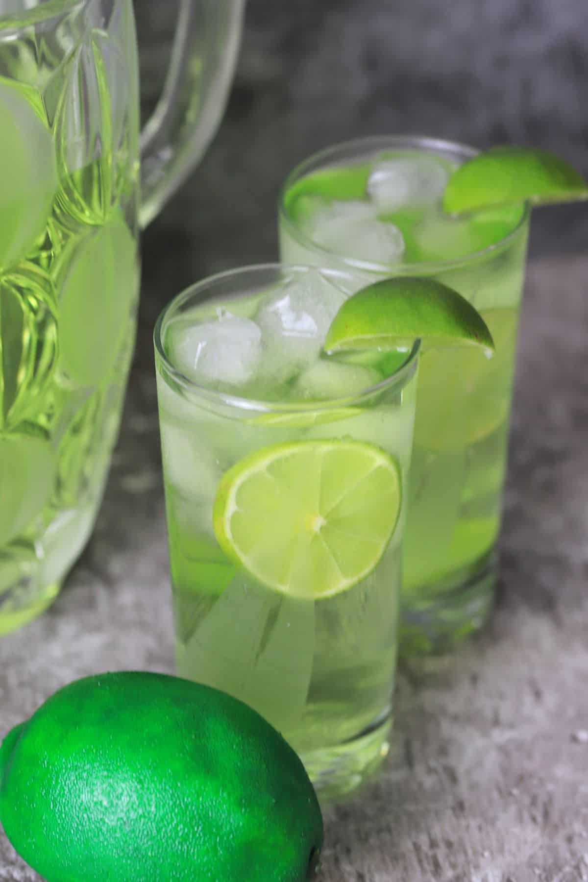 two glasses of the sugar free limeade garnished with lime wedges next to the jug and a whole lime.