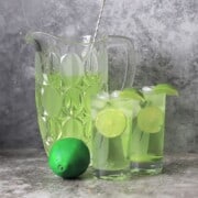 two glasses of the sugar free limeade garnished with lime wedges next to the jug and a whole lime.