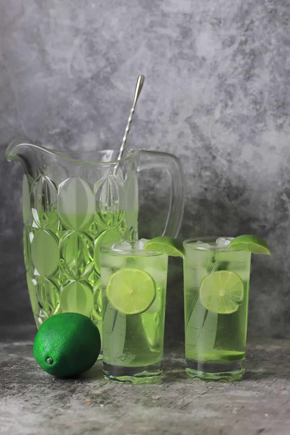 two glasses of the sugar free limeade garnished with lime wedges next to the jug and a whole lime.
