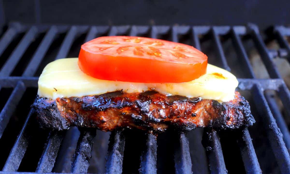 Steak on the grill topped with mozzarella and a tomato slice.