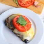 caprese steak on a white plate next to a cutting board with tomatoes and garlic.