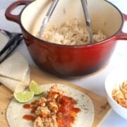 pulled chicken in the dutch oven pot next to a bowl with chicken and taco seasoning and a taco with lime wedges.