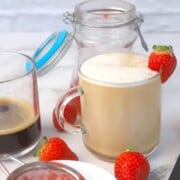 strawberry latte garnished with a strawberry next to a jar, glass with coffee and a strainer.