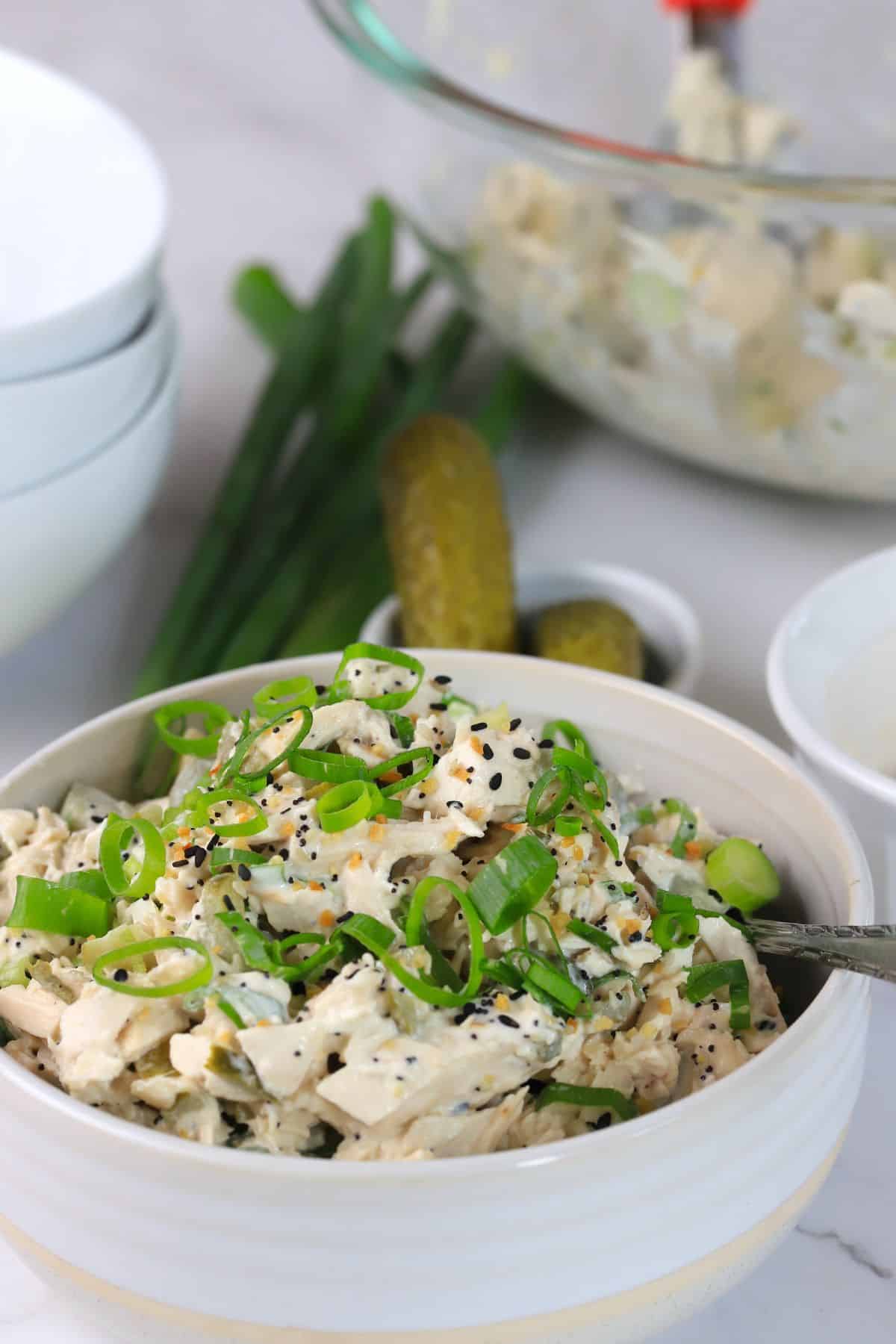 white bowl with chicken salad topped with sliced green onions next to pickles and green onions.