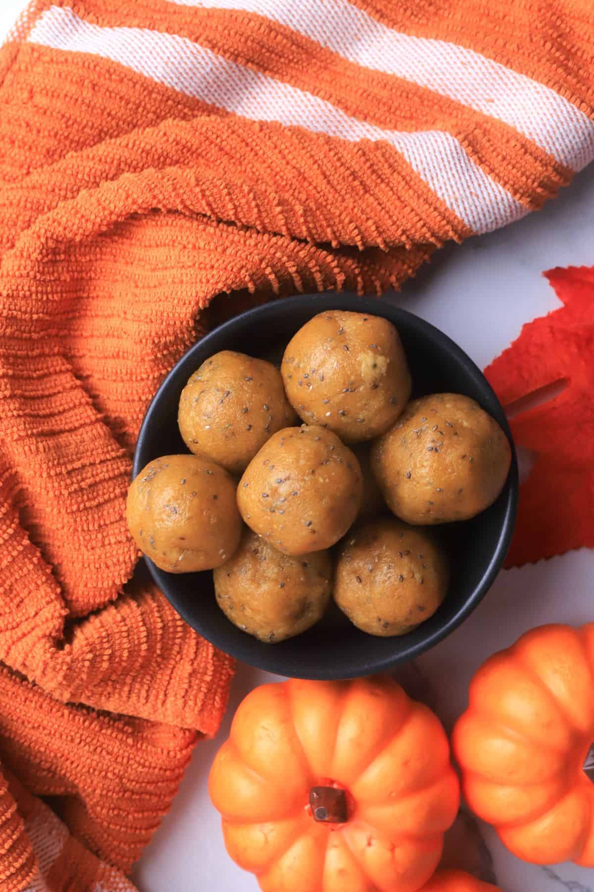 A bowl of pumpkin protein balls in a black bowl surrounded by small pumpkins, an orange leaf and towel.
