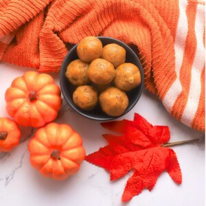 A bowl of pumpkin protein balls in a black bowl surrounded by small pumpkins, an orange leaf and towel.