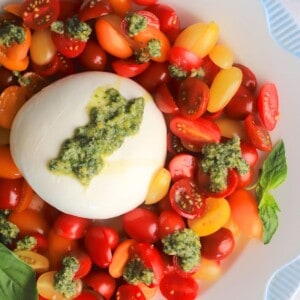 A burrata on a plate surrounded by sliced cherry tomatoes, with dollops of basil pesto and a garnish of basil leaves.