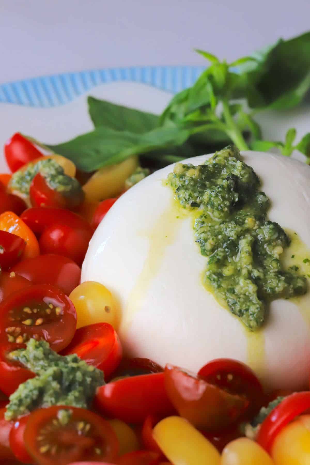 A burrata on a plate surrounded by sliced cherry tomatoes, with dollops of basil pesto and a garnish of basil leaves.