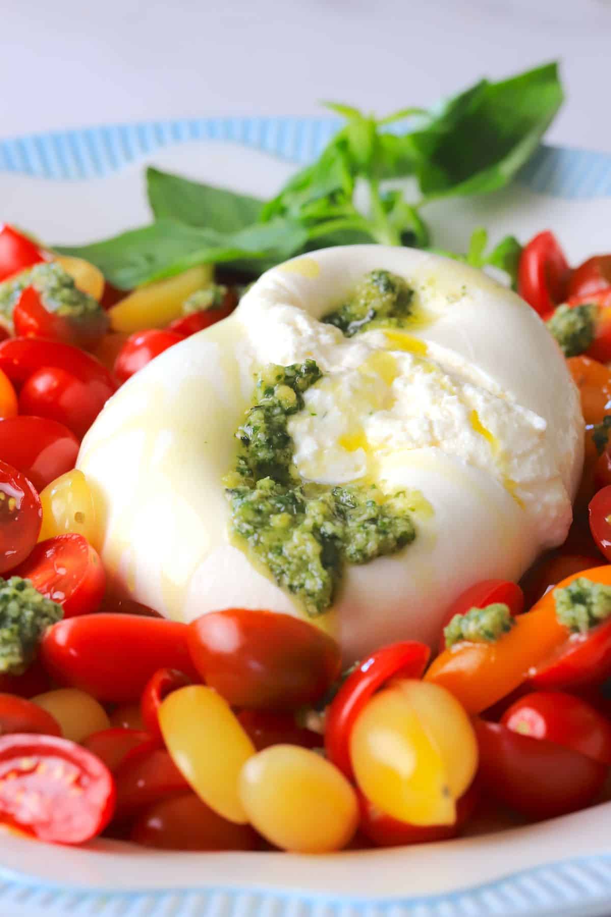 A burrata on a plate surrounded by sliced cherry tomatoes, with dollops of basil pesto and a garnish of basil leaves.