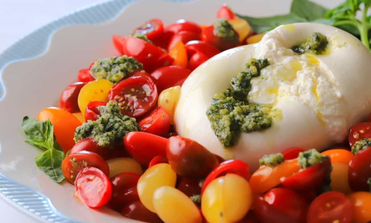 A burrata on a plate surrounded by sliced cherry tomatoes, with dollops of basil pesto and a garnish of basil leaves.