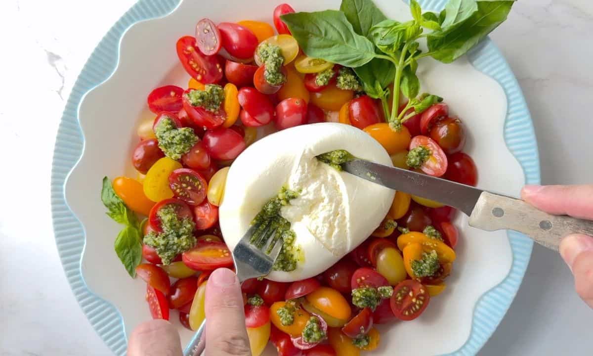 A burrata on a plate surrounded by sliced cherry tomatoes, with dollops of basil pesto and a garnish of basil leaves.