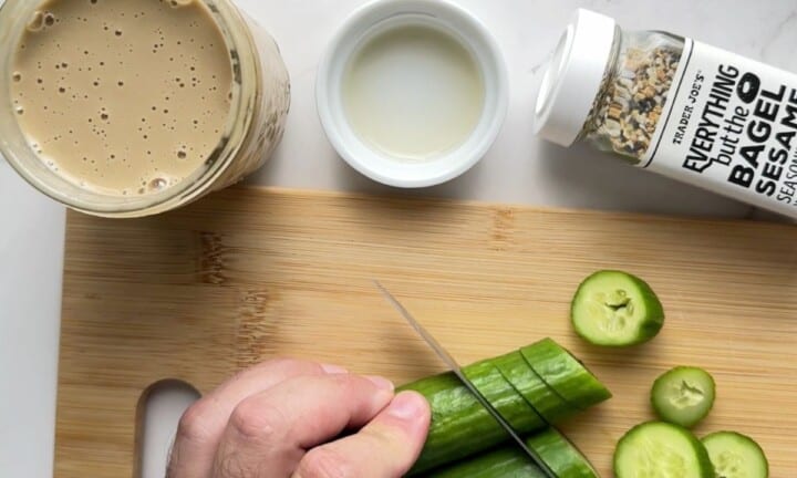 slicing the cucumbers on a cutting board next to the jar of roasted sesame dressing, a small bowl of apple cider vinegar and a jar of everything but the bagel seasoning.
