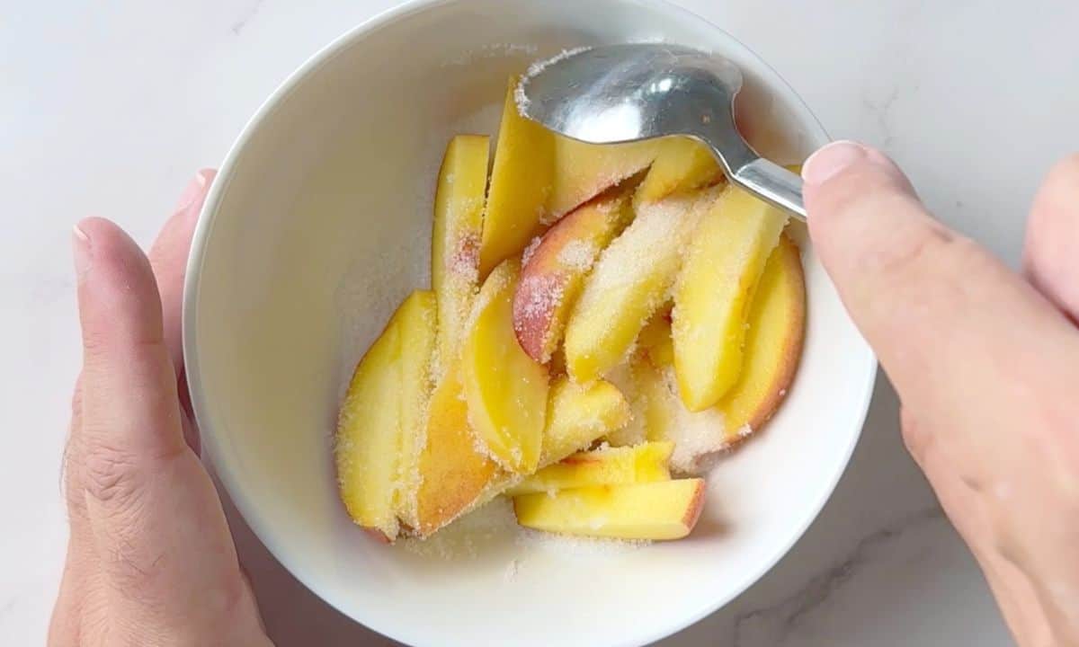 Slices of peaches in a white bowl with sprinkles of granular sweetener and a spoon.