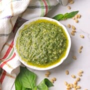 A small bowl of pesto di basilico next to a napkin, some basil leaves and pine nuts.