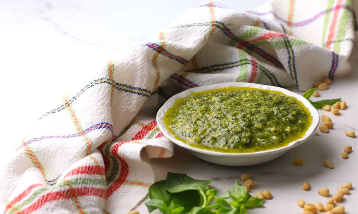 A small bowl of pesto di basilico next to a napkin, some basil leaves and pine nuts.