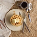 stack of protein powder waffles on a plate with a knife and fork next to a jar with syrup.