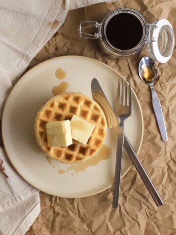 stack of protein powder waffles on a plate with a knife and fork next to a jar with syrup.