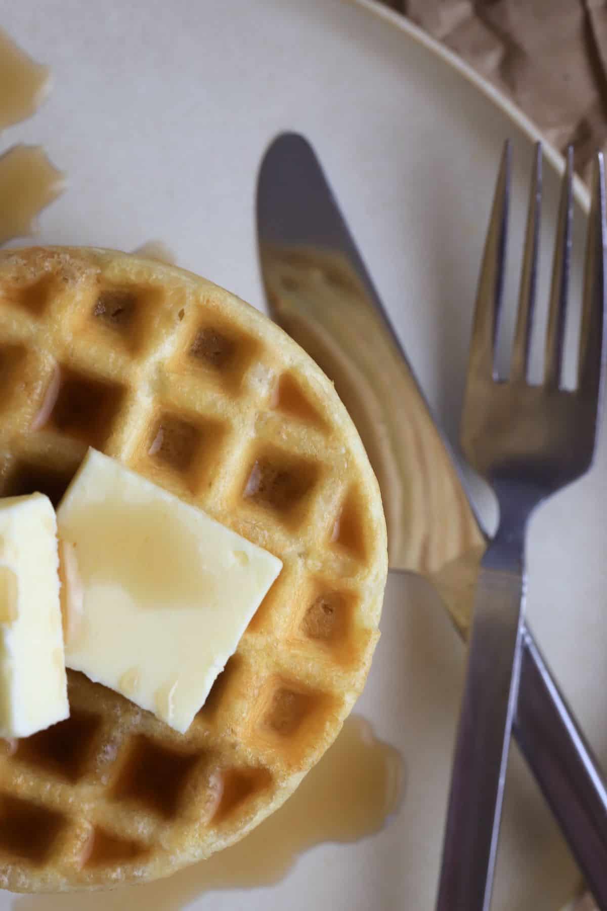 stack of protein powder waffles on a plate with a knife and fork next to a jar with syrup.