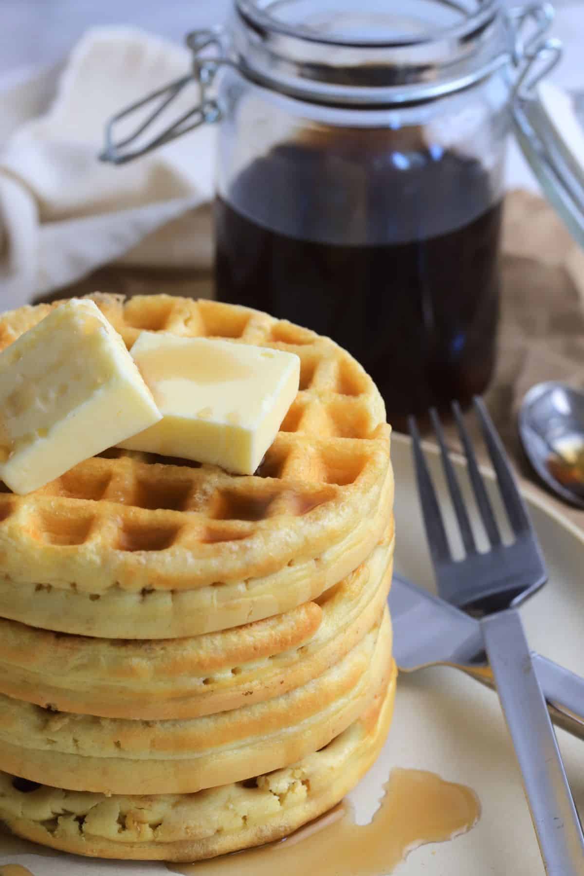 stack of protein powder waffles on a plate with a knife and fork next to a jar with syrup.