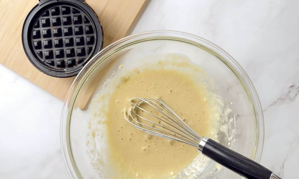 waffles batter in a bowl with a whisk next to the waffle iron.