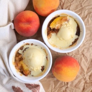 Two peach cobblers in individual ramekins topped with vanilla ice cream and surrounded by 3 fresh peaches.