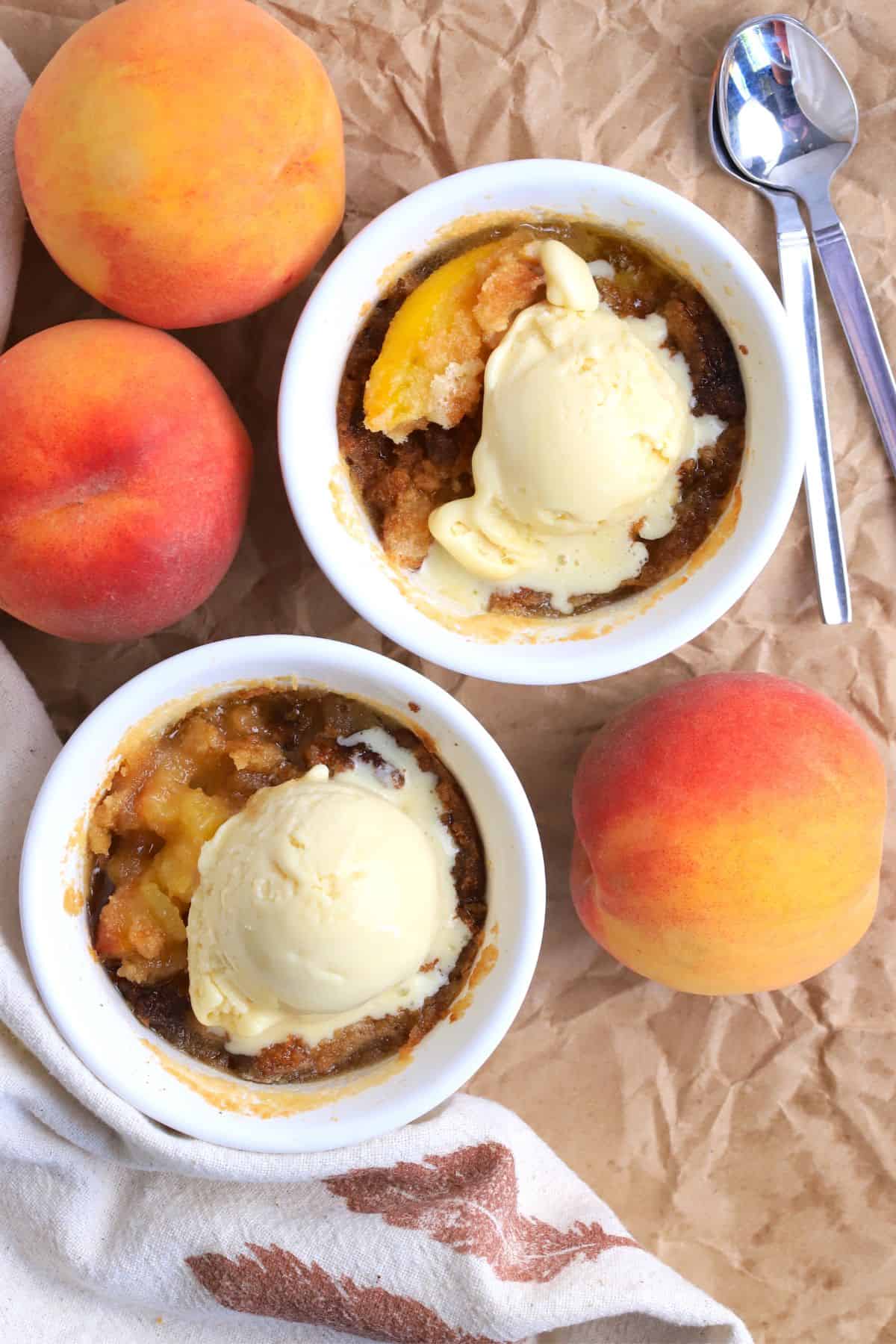 Two peach cobblers in individual ramekins topped with vanilla ice cream and surrounded by 3 fresh peaches.