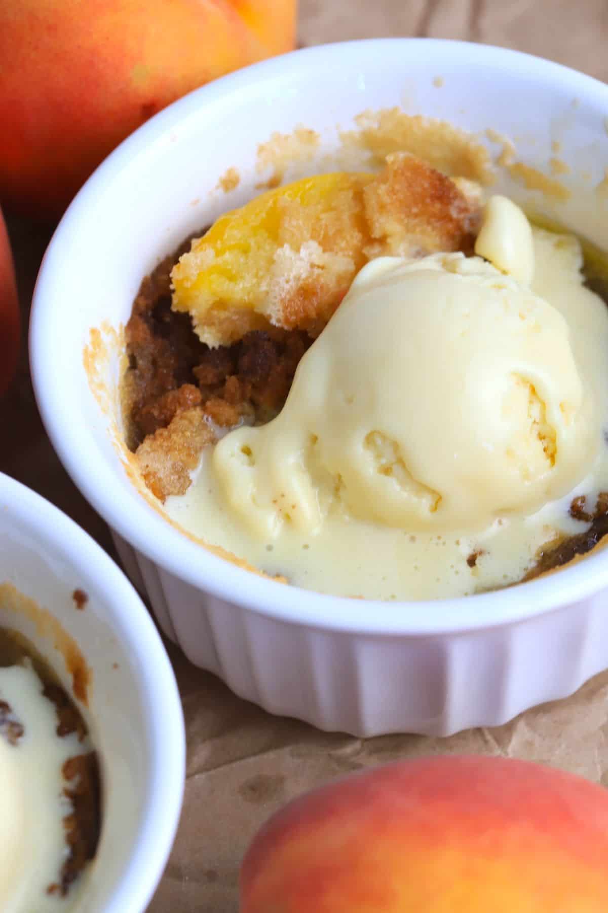 Two peach cobblers in individual ramekins topped with vanilla ice cream and surrounded by 3 fresh peaches.