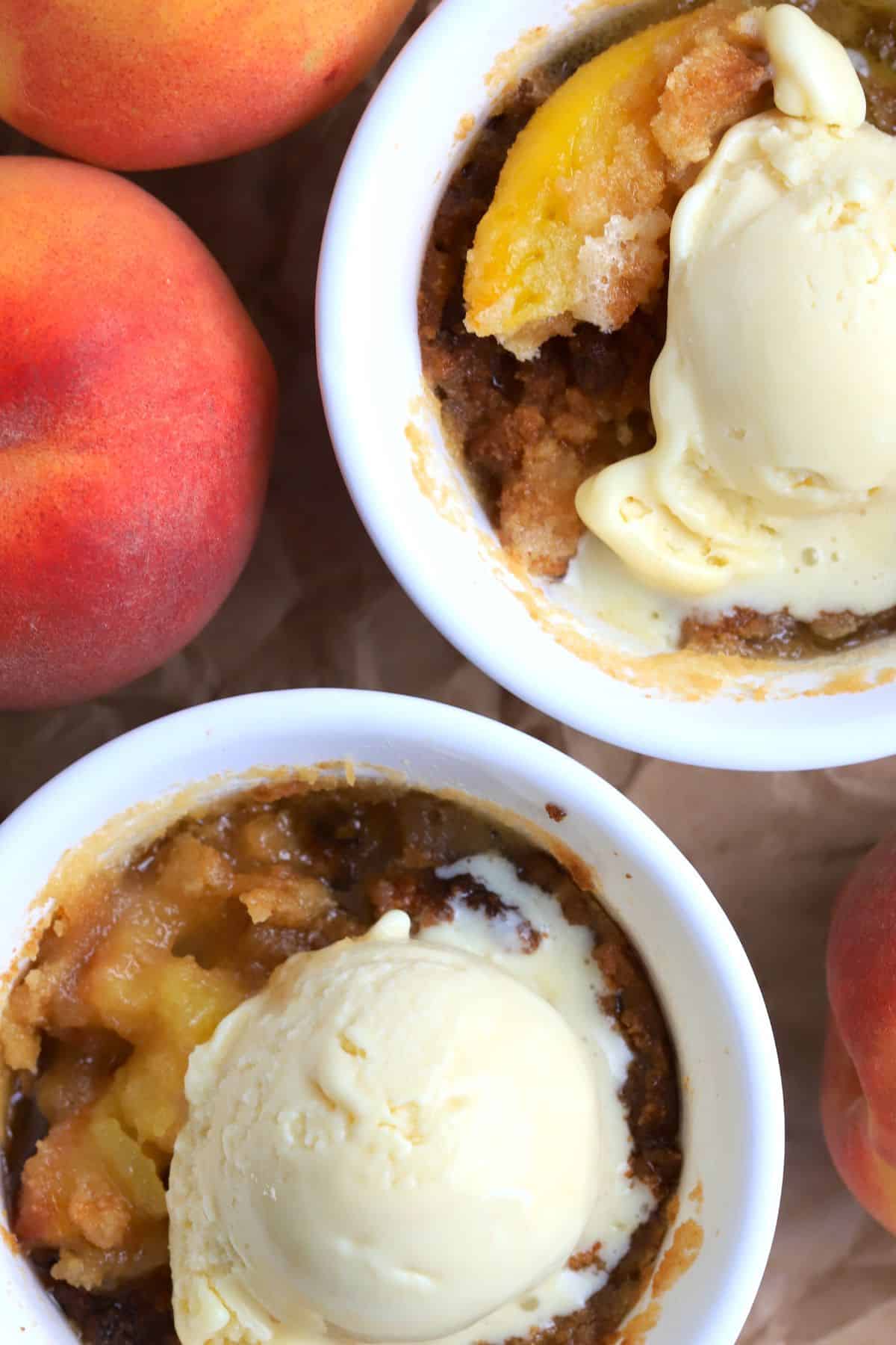 Two peach cobblers in individual ramekins topped with vanilla ice cream and surrounded by 3 fresh peaches.