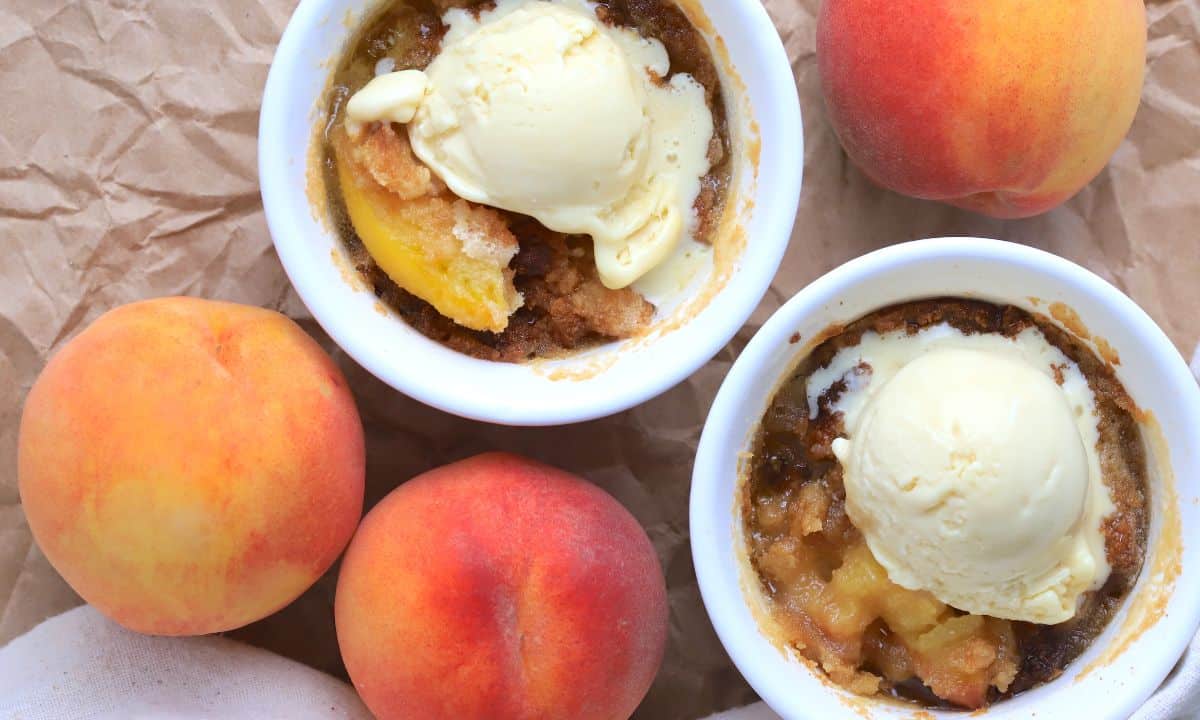 Two peach cobblers in individual ramekins topped with vanilla ice cream and surrounded by 3 fresh peaches.