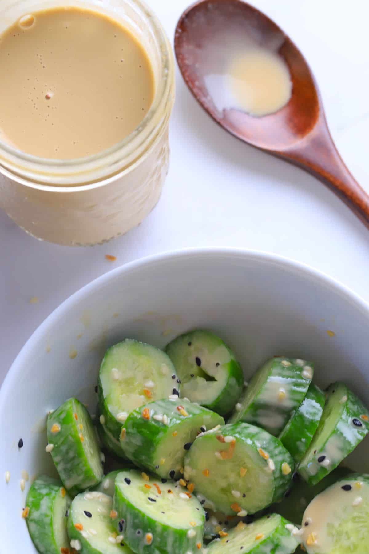 Sugar free salad dressing in a jar next to a wooden spoon and a bowl of sliced cucumbers drizzled with the dressing.