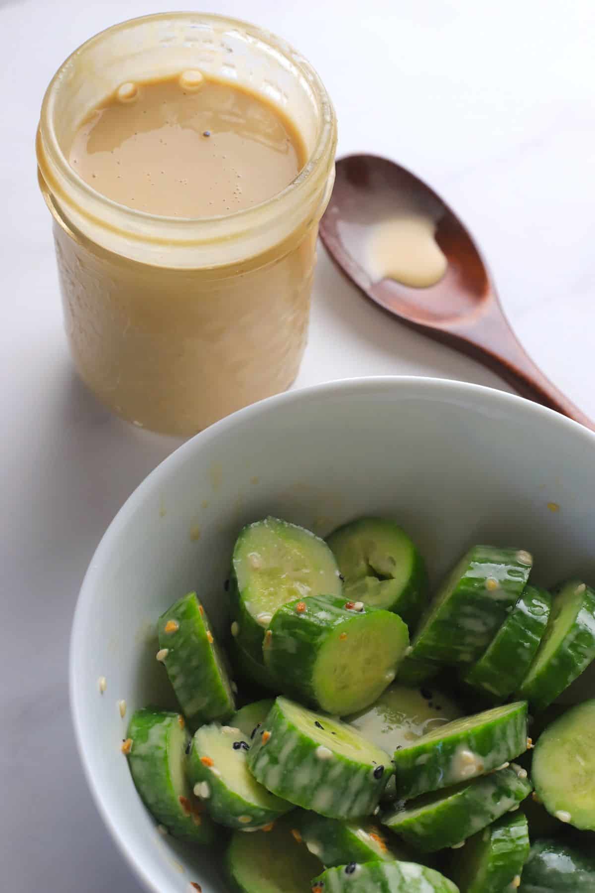 Sugar free salad dressing in a jar next to a wooden spoon and a bowl of sliced cucumbers drizzled with the dressing.