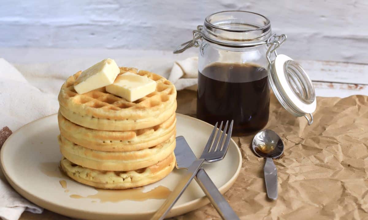 stack of protein powder waffles on a plate with a knife and fork next to a jar with syrup.